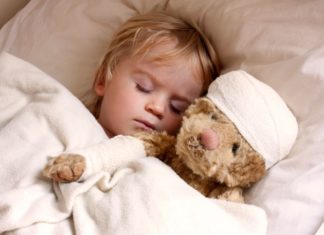 A boy is in bed with his teddy bear. The bear has a bandage around its head.