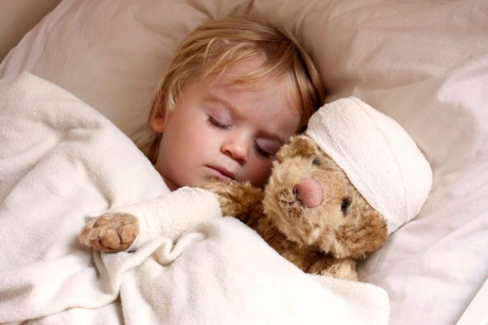 A boy is in bed with his teddy bear. The bear has a bandage around its head.