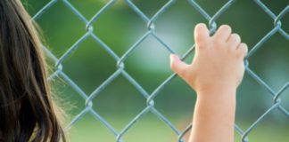 Rear view of a girl standing against a fence, facing a field.