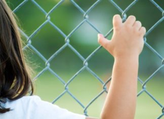 Rear view of a girl standing against a fence, facing a field.