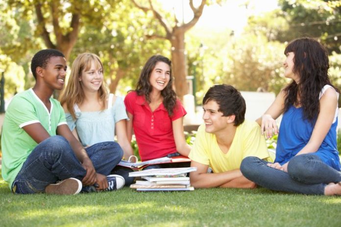 An eclectic and picture perfect group of school kids hang out on the lawn in front of a pile of books.