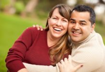 A couple hug and smile to camera. The man is wearing a ribbed turtle neck.