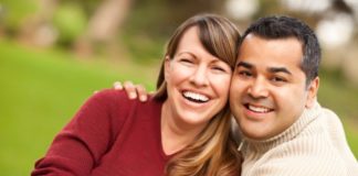 A couple hug and smile to camera. The man is wearing a ribbed turtle neck.