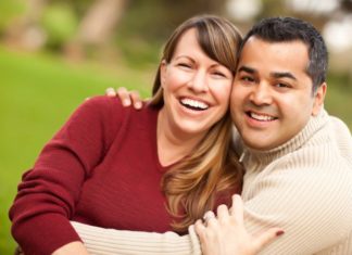 A couple hug and smile to camera. The man is wearing a ribbed turtle neck.