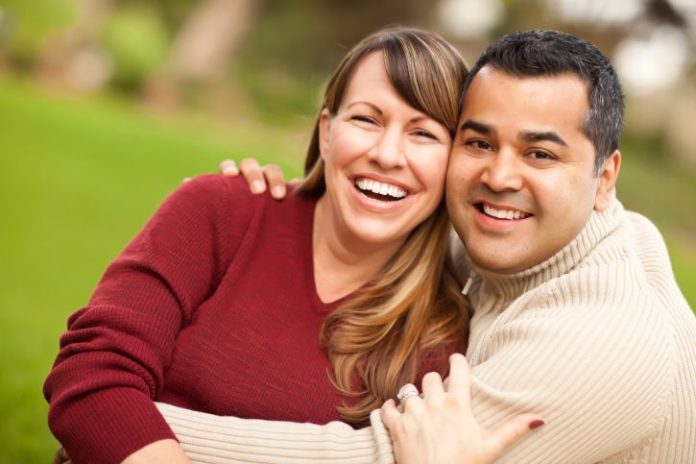 A couple hug and smile to camera. The man is wearing a ribbed turtle neck.