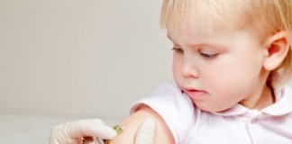 A little girl in pink looks at her arm as a gloved hand gives her an injection. For a toddler, she is very well composed.