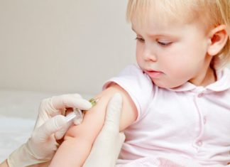 A little girl in pink looks at her arm as a gloved hand gives her an injection. For a toddler, she is very well composed.