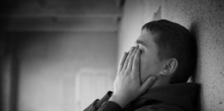 Teenager leans against wall, covering face with hands.