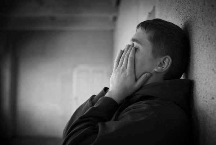 Teenager leans against wall, covering face with hands.