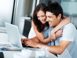 A thrilled looking couple sit at a table using a laptop. The woman has her arms around the man's shoulders.