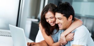 A thrilled looking couple sit at a table using a laptop. The woman has her arms around the man's shoulders.