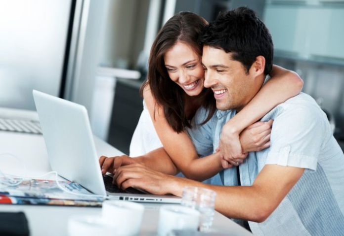 A thrilled looking couple sit at a table using a laptop. The woman has her arms around the man's shoulders.