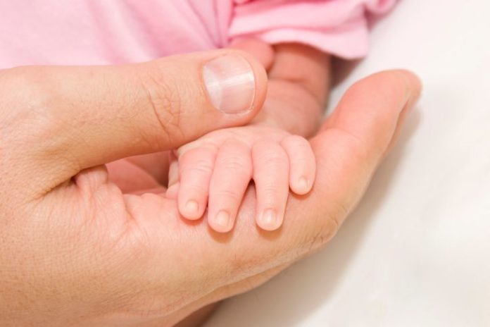 A very small baby's hand sits in the hand of her father.