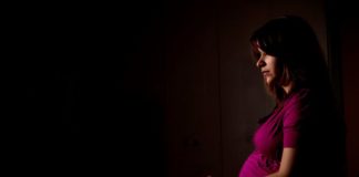 Pregnant woman in a dark scene. She stares ahead blankly.