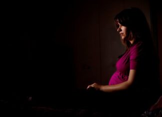 Pregnant woman in a dark scene. She stares ahead blankly.