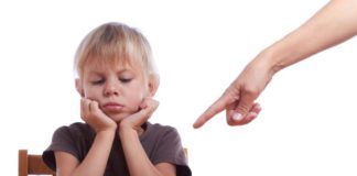 An adult's hand points into the shot at a bowl. The boy sits in front of the bowl looking sullen, holding his chin in his hands.