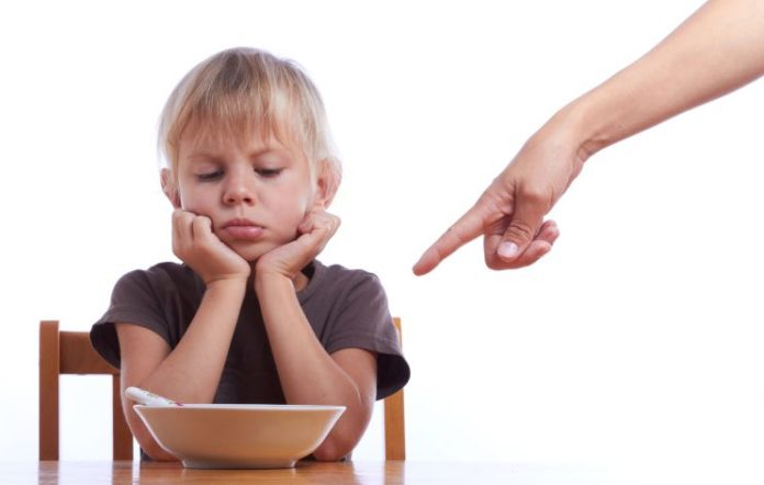 An adult's hand points into the shot at a bowl. The boy sits in front of the bowl looking sullen, holding his chin in his hands.