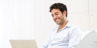 A man sits on a couch smiling, using a generic laptop.