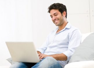 A man sits on a couch smiling, using a generic laptop.