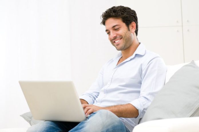 A man sits on a couch smiling, using a generic laptop.