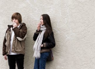 A teen girl and boy lean against a wall smoking.