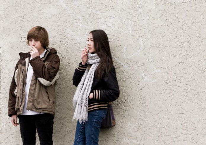 A teen girl and boy lean against a wall smoking.