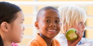 Three children sit together. One boy smiles to camera and shows his green apple that he has been eating.