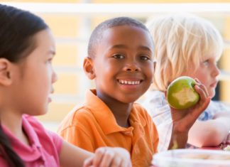 Three children sit together. One boy smiles to camera and shows his green apple that he has been eating.