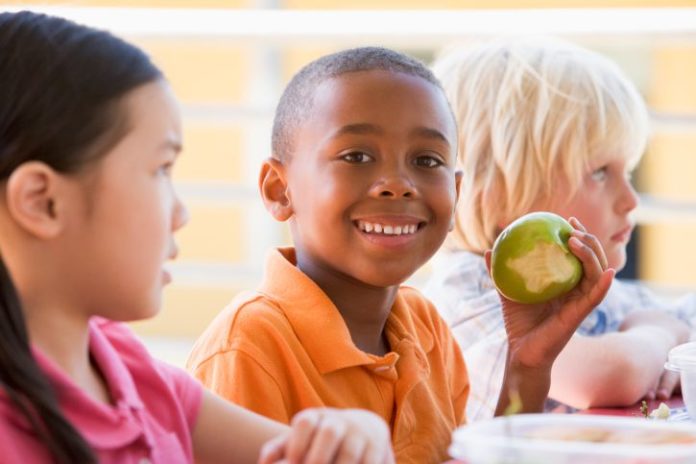 Three children sit together. One boy smiles to camera and shows his green apple that he has been eating.