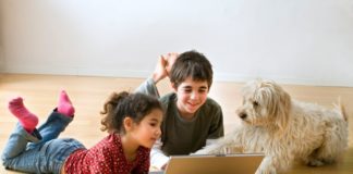 Two kids using a laptop lay on the ground with their dog.