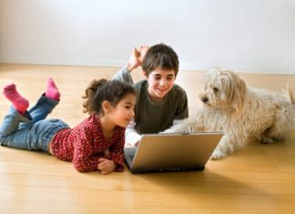 Two kids using a laptop lay on the ground with their dog.