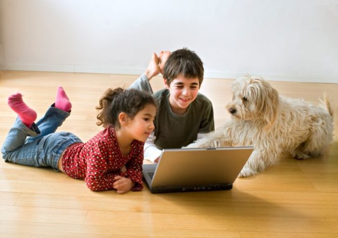 Two kids using a laptop lay on the ground with their dog.