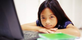 A girl sits in front of her laptop. Her chin rests on her hands. She is unhappy.