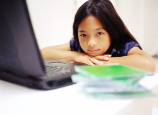 A girl sits in front of her laptop. Her chin rests on her hands. She is unhappy.