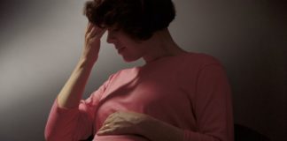 Pregnant woman in a dark scene. She had her fingers to her forehead and her eyes closed.