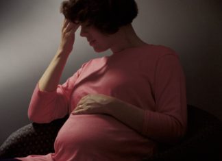 Pregnant woman in a dark scene. She had her fingers to her forehead and her eyes closed.