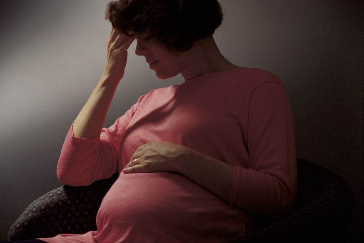 Pregnant woman in a dark scene. She had her fingers to her forehead and her eyes closed.