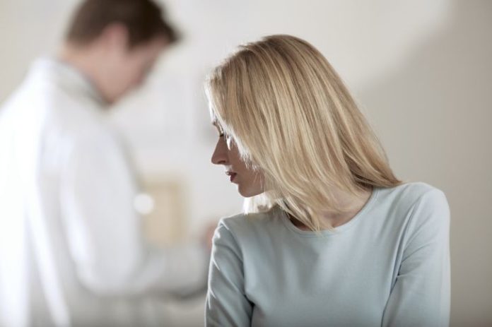 A depressed looking scene as a woman looks over her shoulder the the doctor in the background. He is out of focus.