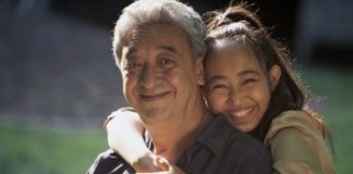 Daughter hugs father around the neck. Both smile to camera.