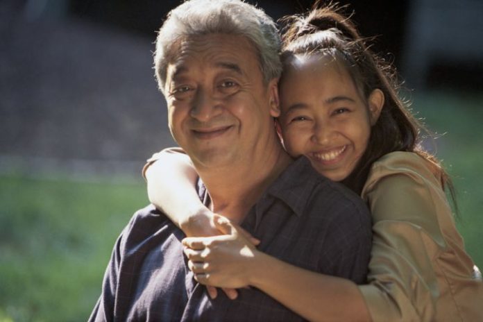 Daughter hugs father around the neck. Both smile to camera.