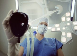 A doctor in scrubs holds a mask to be placed over the patient. The patient is the camera.