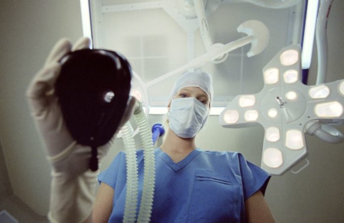 A doctor in scrubs holds a mask to be placed over the patient. The patient is the camera.