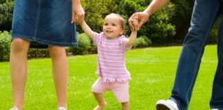 Girl walking with parents on grass, holding both parents' hands.