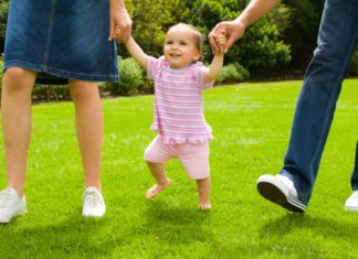 Girl walking with parents on grass, holding both parents' hands.