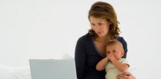 A mother sits on her bed holding her baby. She works on a laptop at the same time.