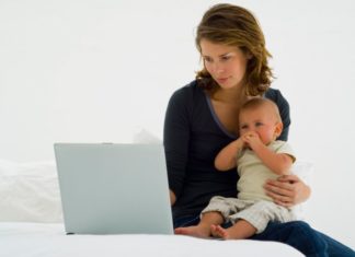 A mother sits on her bed holding her baby. She works on a laptop at the same time.
