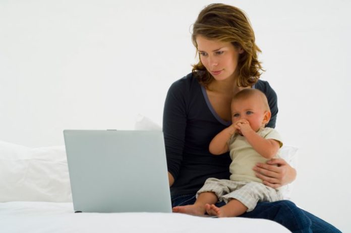 A mother sits on her bed holding her baby. She works on a laptop at the same time.