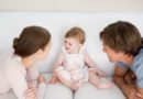 A baby sits on the couch as the two parents huddle in front.