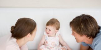 A baby sits on the couch as the two parents huddle in front.