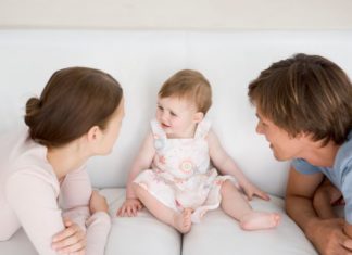 A baby sits on the couch as the two parents huddle in front.